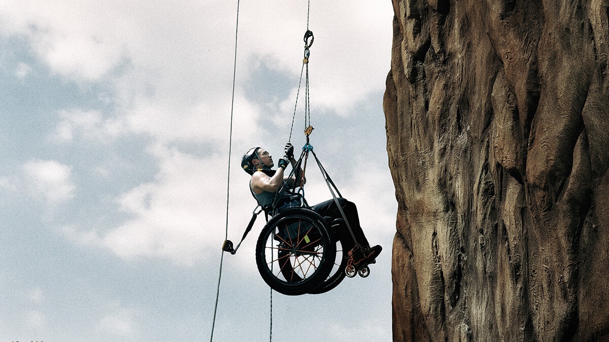 A climber hoists himself by pulling ropes attached to his body and wheelchair up along a rocky vertical cliff.