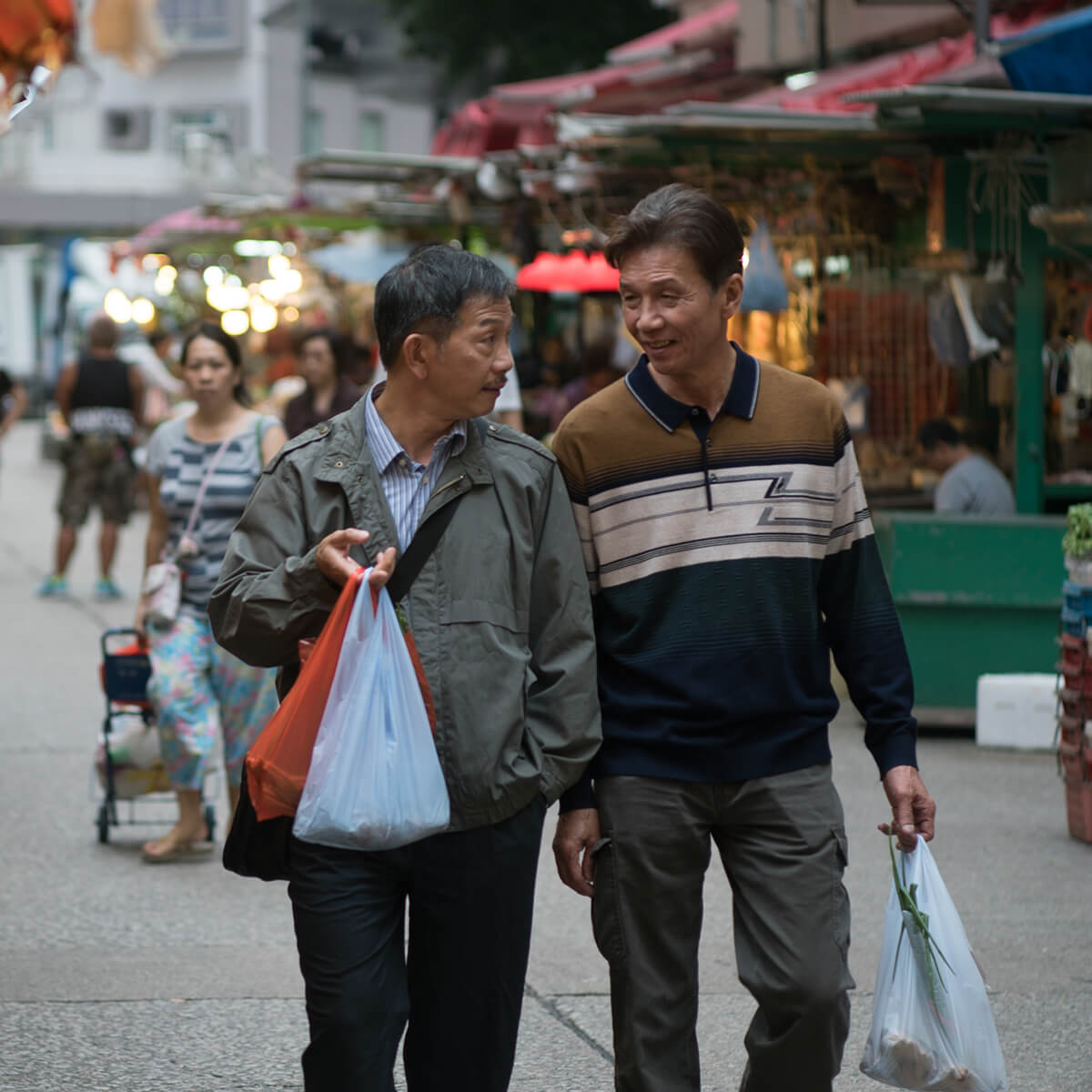 Two men holding shopping bags chat and walk through an outdoor marketplace.