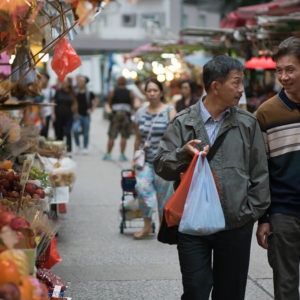 Two men holding shopping bags chat and walk through an outdoor marketplace.