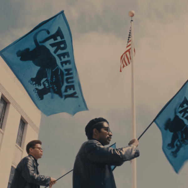Two men wave black panther flags in front of white building under a blue sky