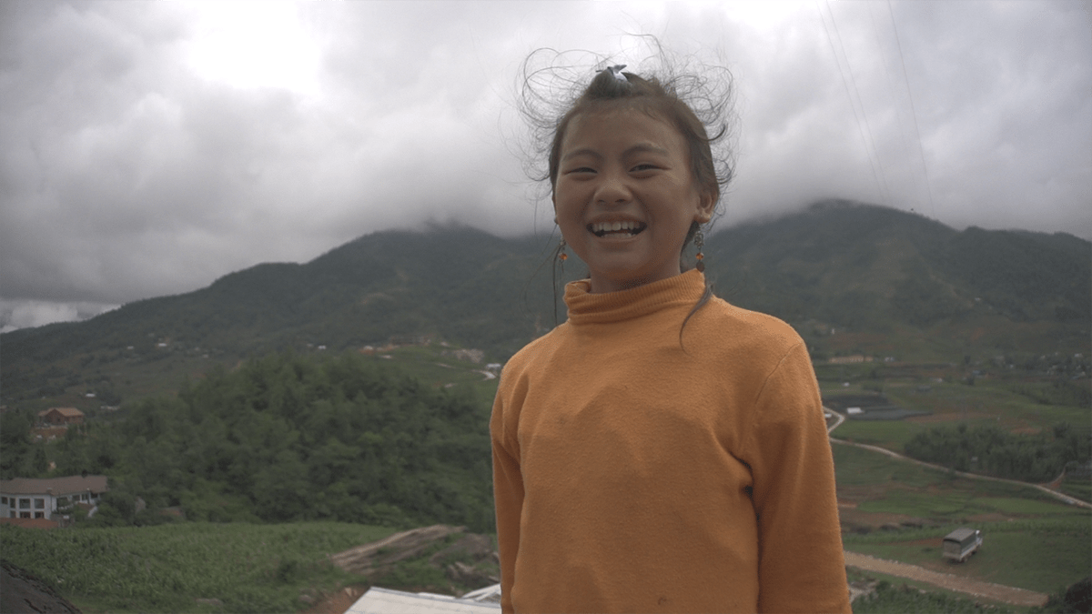 a child whose hair is being blown by the wind stands in front of mountains