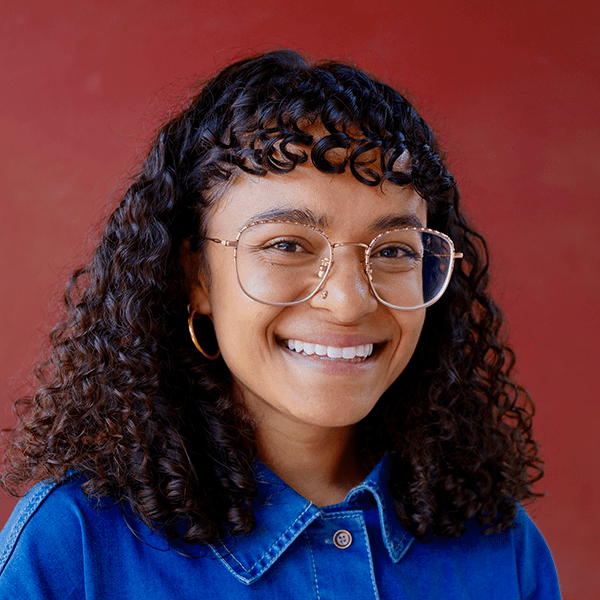 person with curly hair and glasses smiling in front of burgundy background