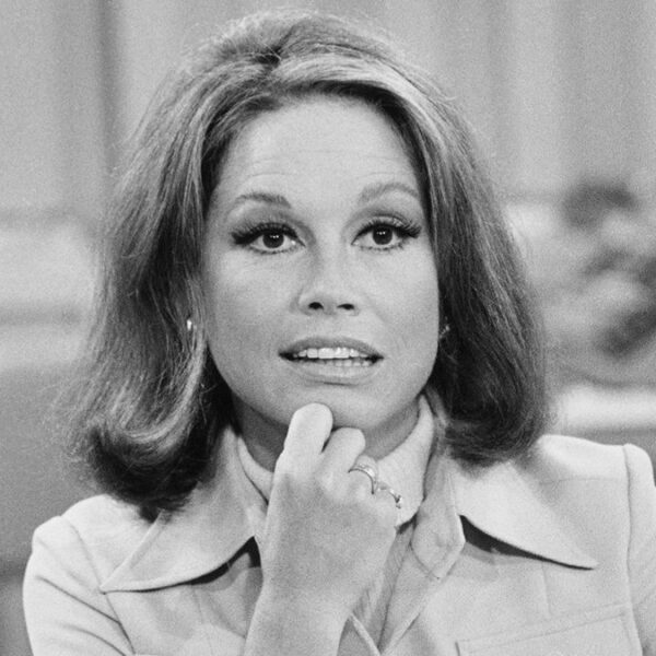a black and white photo of a woman wearing a suit and turtle neck sitting at a desk.