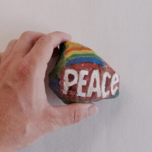 a hand holding a multi colored painted rock with the word "Peace" on it.