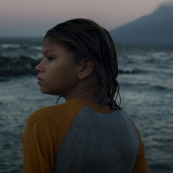 a young girl standing by a body of water with a mountain in the distance.