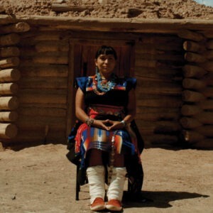 a woman in native dress sitting in front of a wooden house.