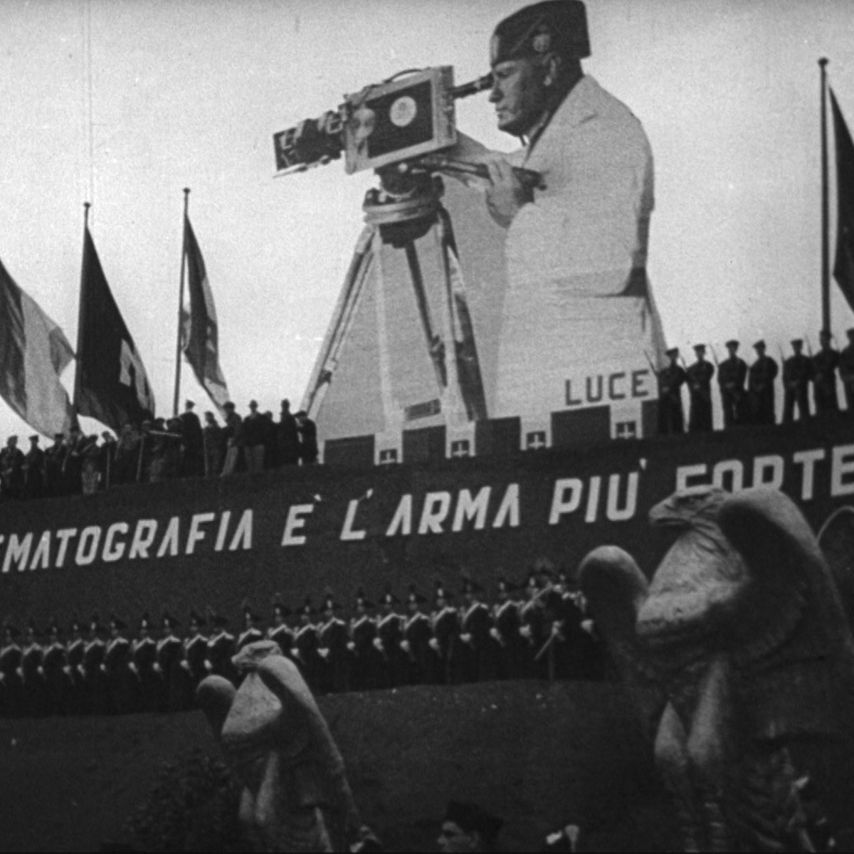 a large cutout of a man holding a camera stands over a crowd of soldiers.
