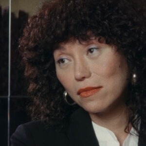 a woman with curly hair and red lipstick sitting by a mirrored wall.