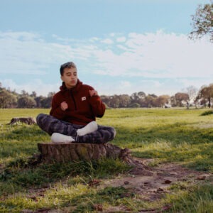 a young person sitting cross legged on a tree stump in a field of green grass.