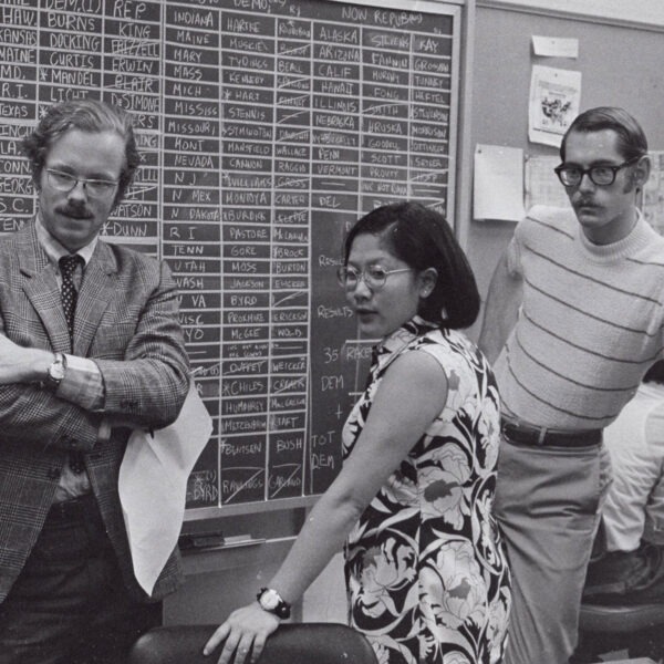 a black and white photo of two men and a woman standing in front of a chalkboard marked up with names.
