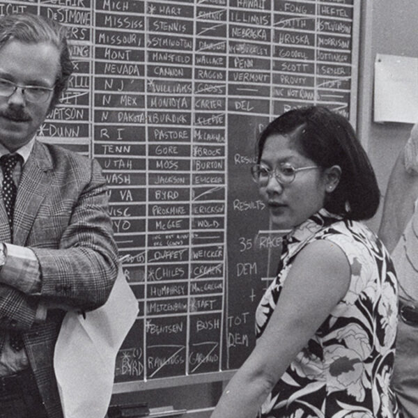 a black and white photo of two men and a woman standing in front of a chalkboard marked up with names.