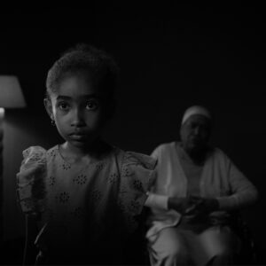 a black and white still of a child standing in front of a woman in a wheelchair by a lamp.