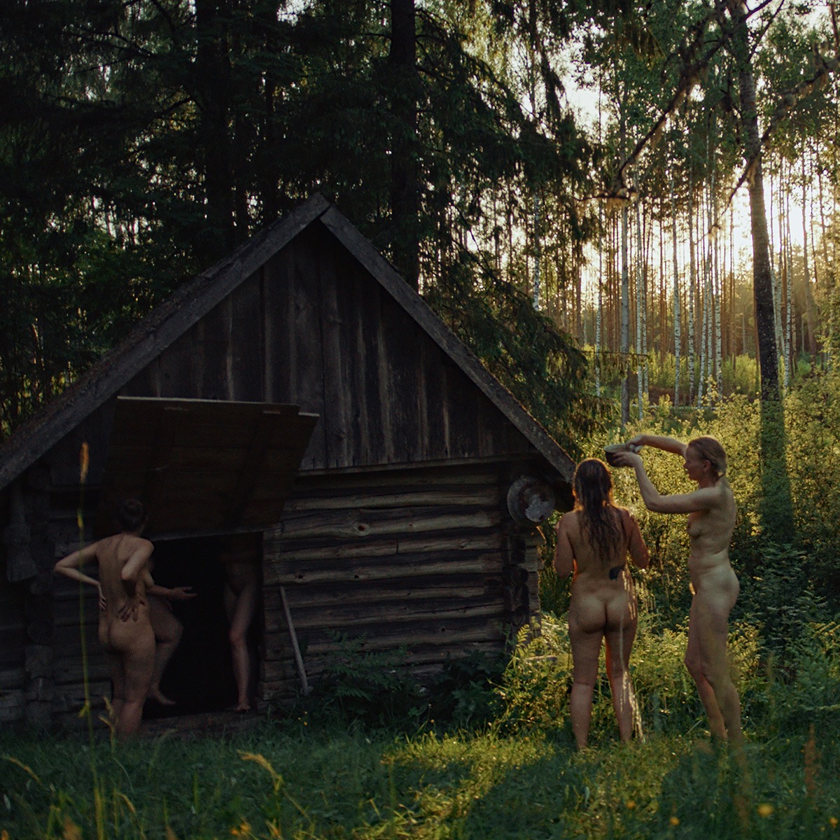 a group of naked woman stand by a wooden hut in a forest.