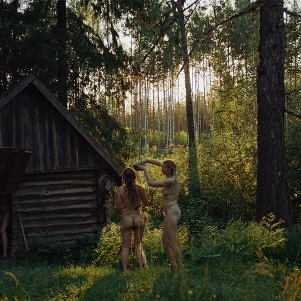 a group of naked woman stand by a wooden hut in a forest.