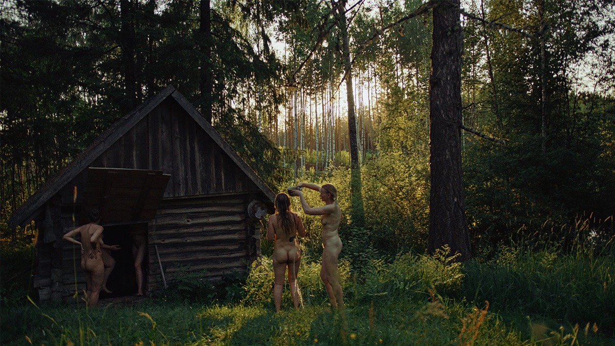a group of naked woman stand by a wooden hut in a forest.