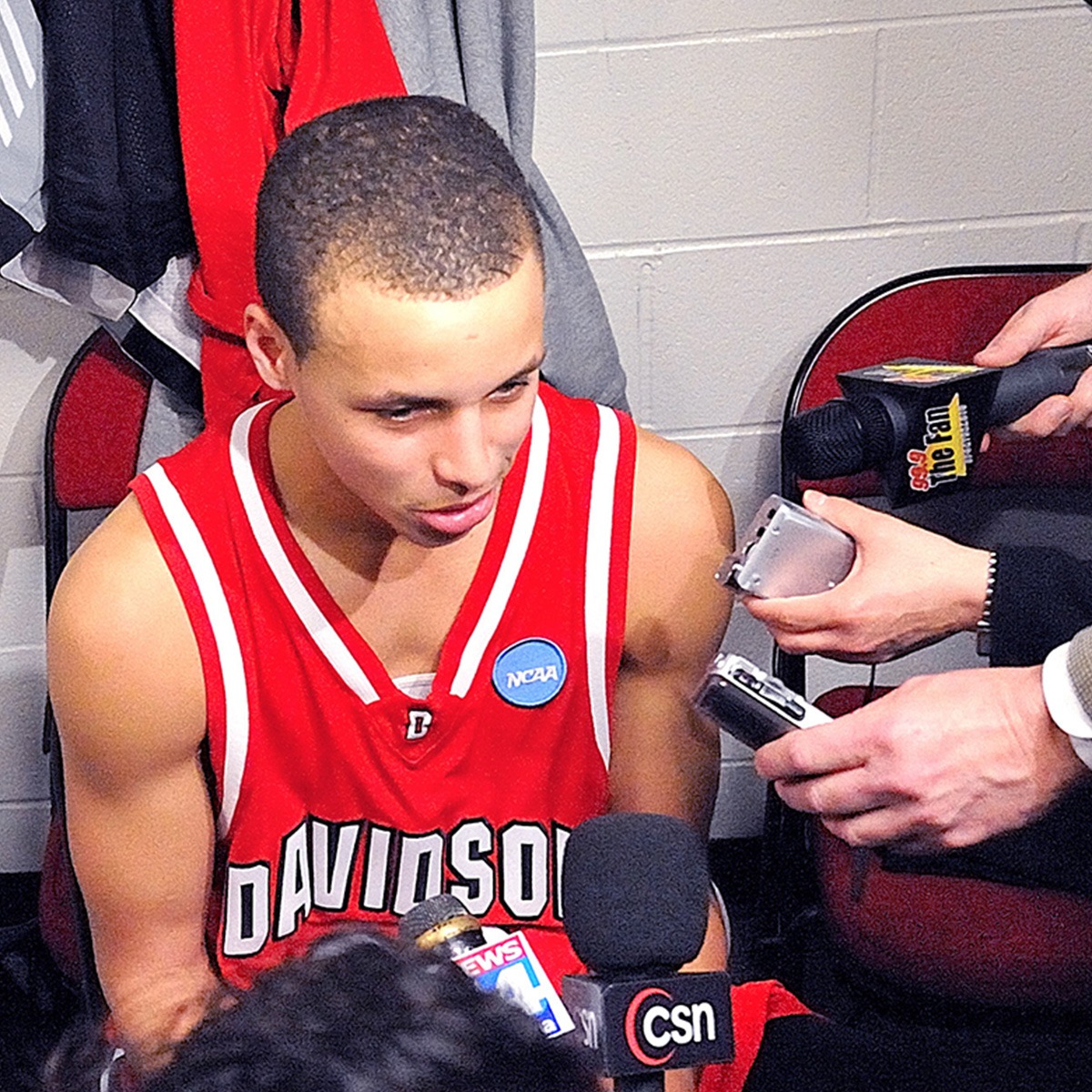 a young stephen curry in a red jersey sitting surrounded by microphones.