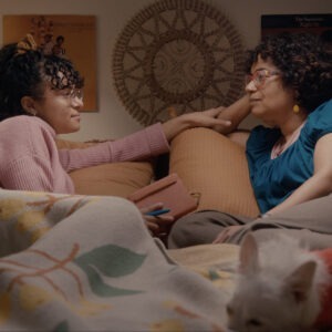 a couple of women face each other sitting on top of a bed.