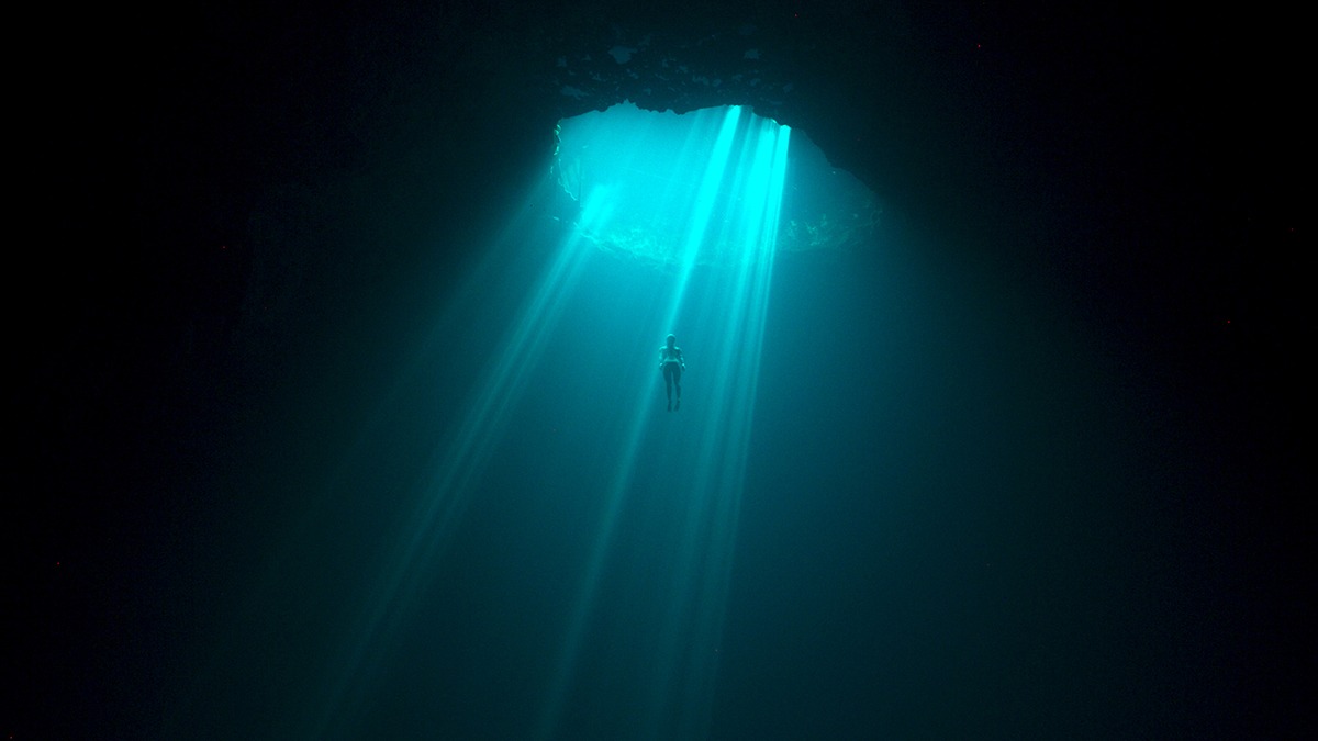 a woman underwater swimming up to a hole that has light shining through.