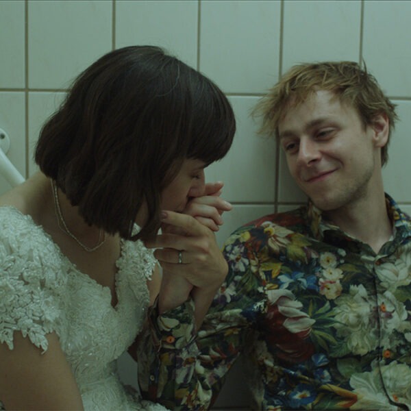 a woman in a white dress kissing a man's hand sit in a restroom.
