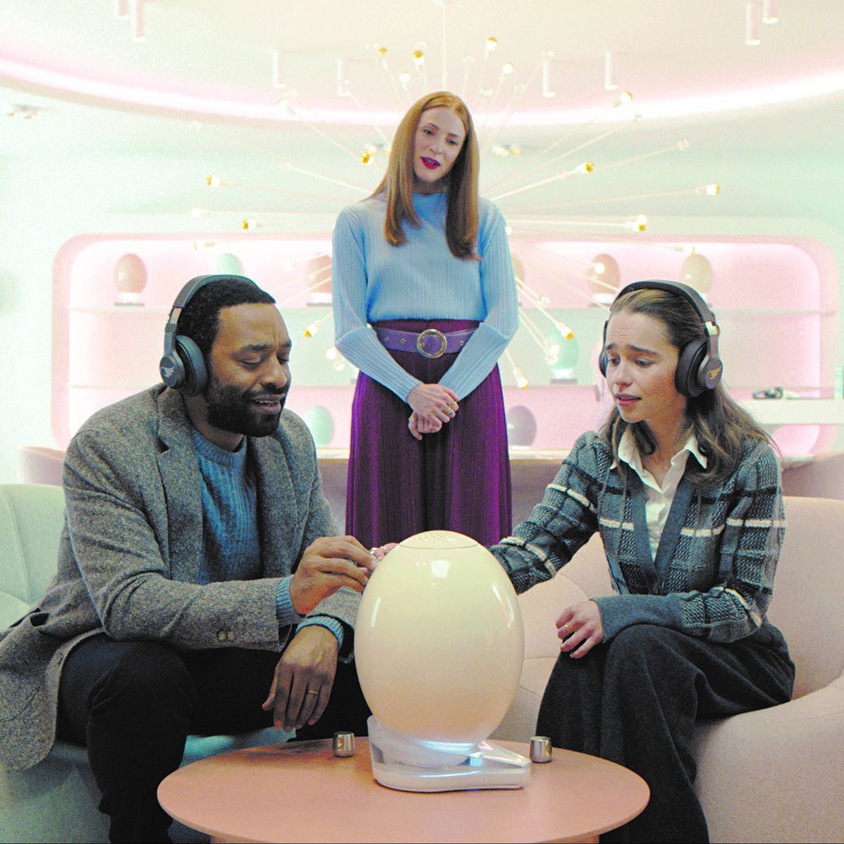 a man and a woman wearing headphones sitting next to a white pod in a futuristic room.