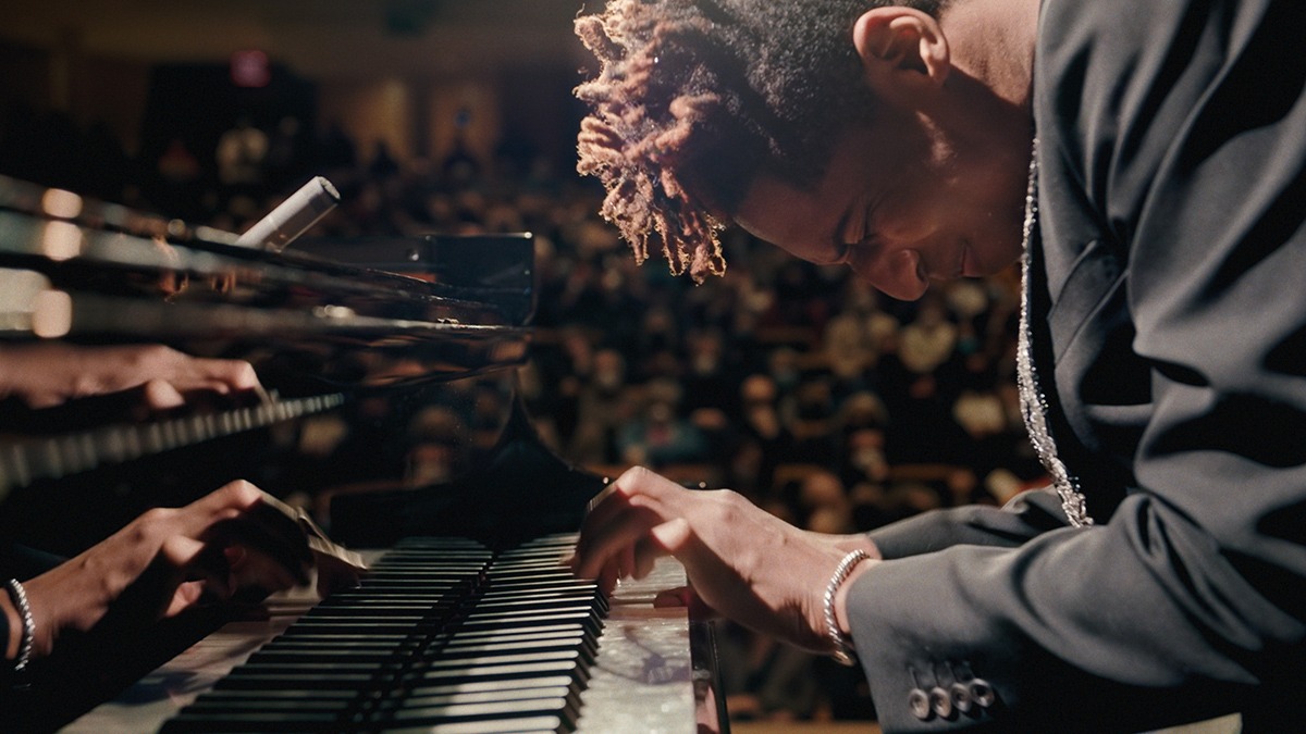 John Batiste, a black man in a suit playing a piano as a large audience watches him.