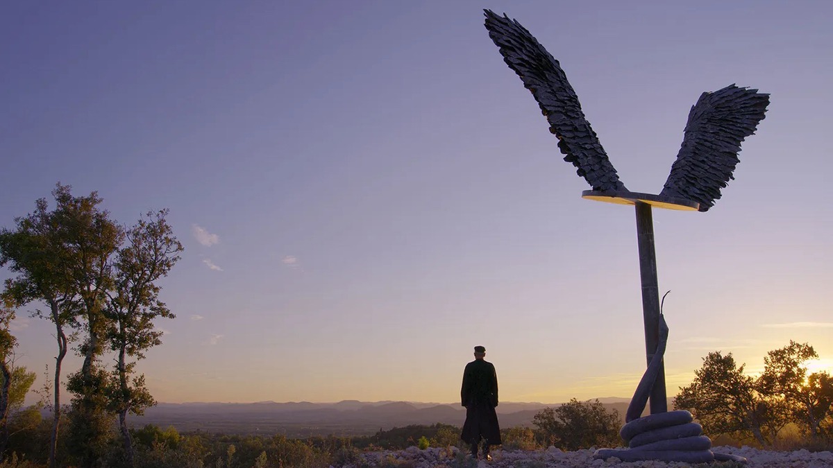 a lone figure standing beside a tall statue with wings watches the sunrise in an open field.