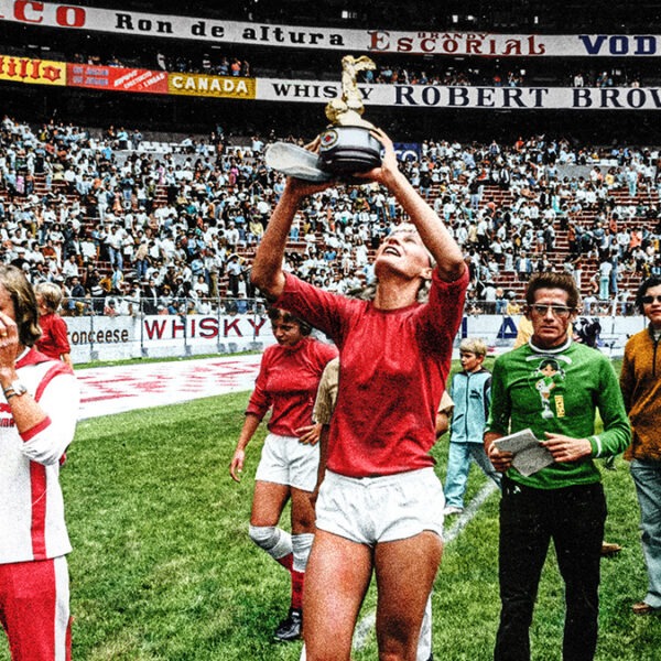 a white woman soccer player holds up a trophy amongst her team in a soccer field full of audience members.