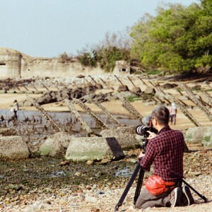 a man kneeling by his camera on a tripod pointing at an old stone war fort by the shore.