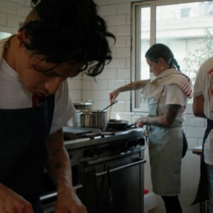 three chefs wearing aprons actively cooking at stoves in a restaurant kitchen.