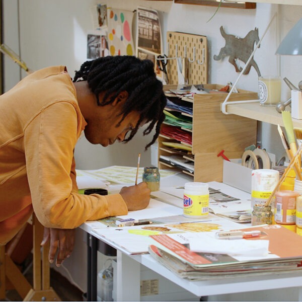 a young black man hunches over a workshop desk sketching.