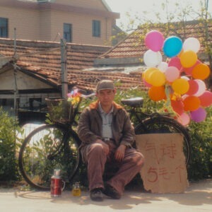 A man sitting in front of a bicycle with a bunch of ballons in his hand.