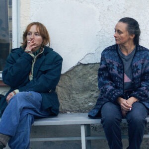Two women sitting on a bench next to a wall.