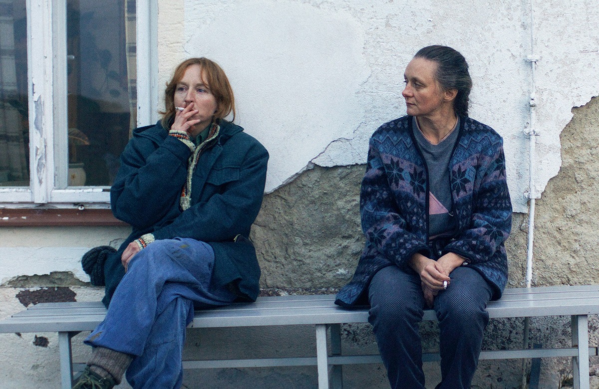 Two women sitting on a bench next to a wall.